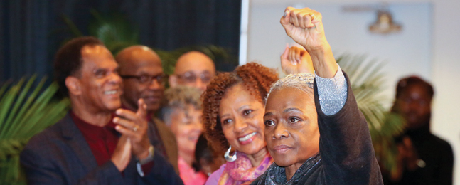 A black woman raises her fist as men and women around her applaud.